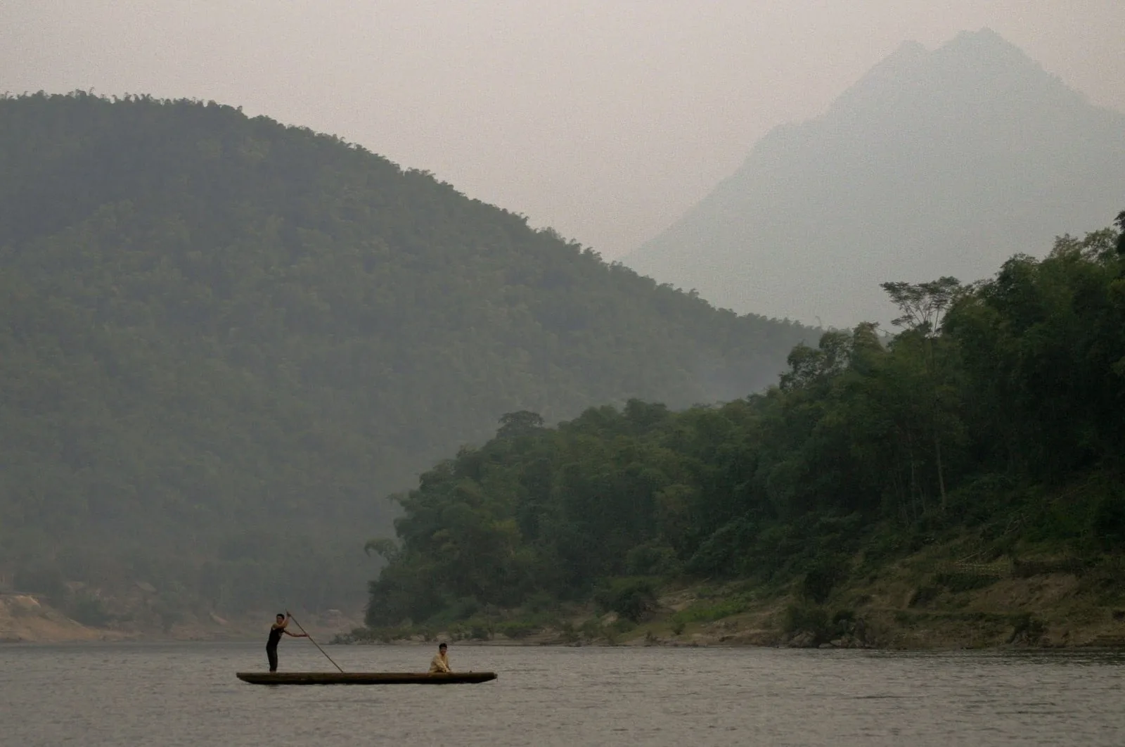 Two people paddle a small wooden boat along a misty river bordered by forested mountains near Mai Chau.