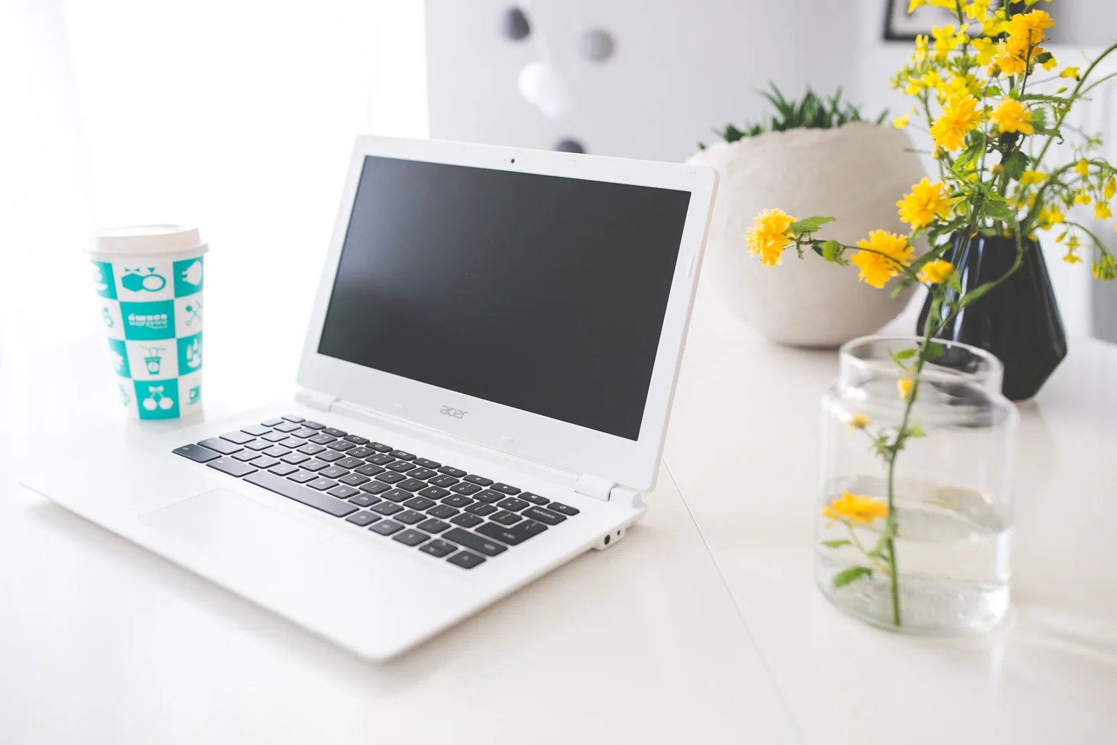 A white laptop sits on a desk beside a coffee cup and yellow flowers in a bright home office.