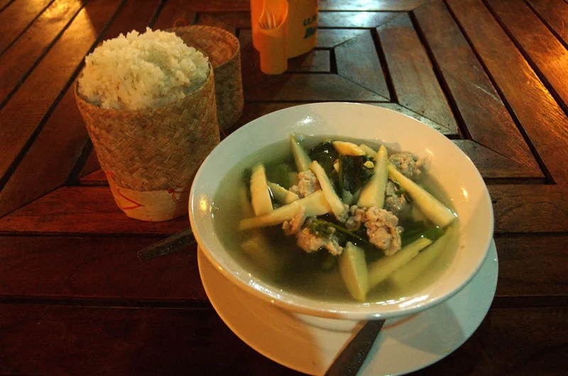 A bowl of bamboo shoot soup with minced meat served beside sticky rice in a woven basket in Luang Namtha.