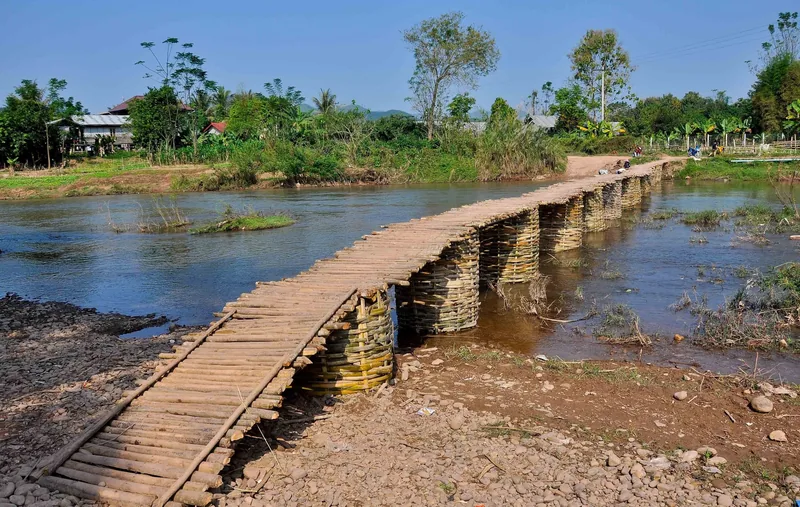 A traditional bamboo bridge crosses a river near Luang Namtha, Laos, under a clear blue sky.