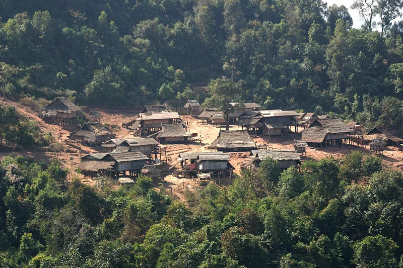 A cluster of stilted wooden houses forms a small hillside village surrounded by dense forest near Luang Namtha.