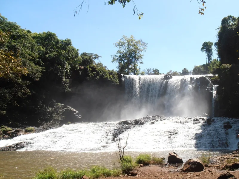 A wide waterfall crashes over dark rocks into a pool, surrounded by dense green forest in Mondulkiri.