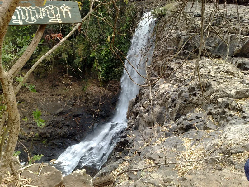 Bou Sra waterfall cascades over dark volcanic rock in Mondulkiri, with a wooden sign in Khmer script nearby.