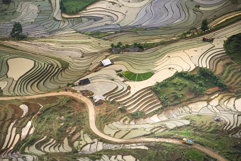 Aerial view of winding terraced rice paddies with scattered farmhouses and a dirt road near Sapa, Vietnam.