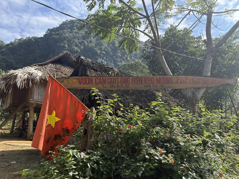 A thatched-roof stilt house with a Vietnamese flag and a hand-painted sign stands amid greenery in Pu Luong.