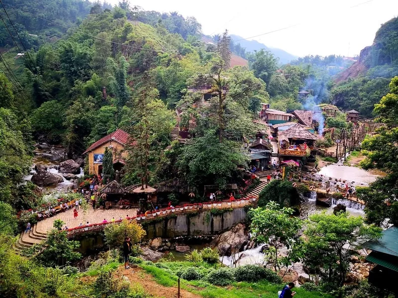 A stone pathway winds through wooden houses and streams surrounded by lush hills in Cat Cat Village, Sapa.