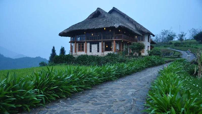 A thatched-roof stone lodge glows with warm light beside a stone path through terraced fields at dusk in Sapa.