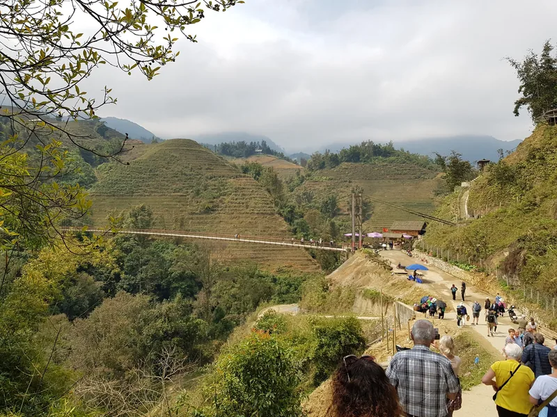 Tourists walk a mountain trail past terraced hills and a suspension bridge near Sapa's Muong Hoa valley.