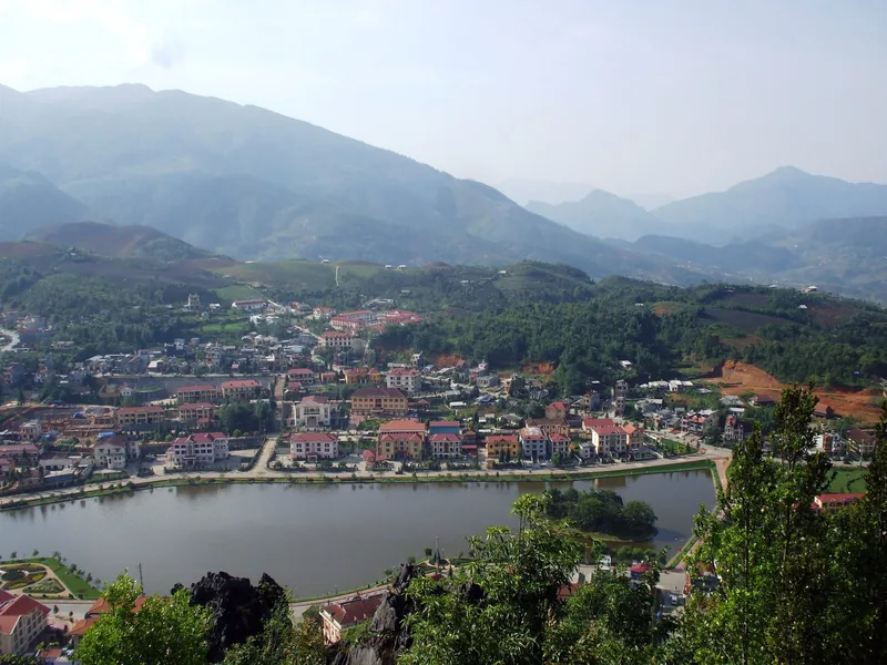 A hillside town in Sapa nestles beside a lake, surrounded by layered mountains and forest.